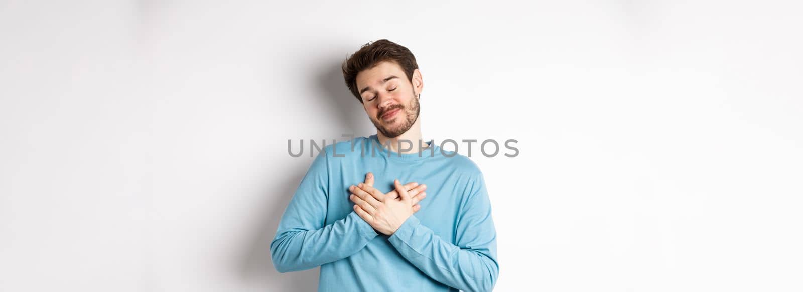 Handsome young man feeling romantic, holding hands on heart and thinking about good pleasant memories, standing over white background.