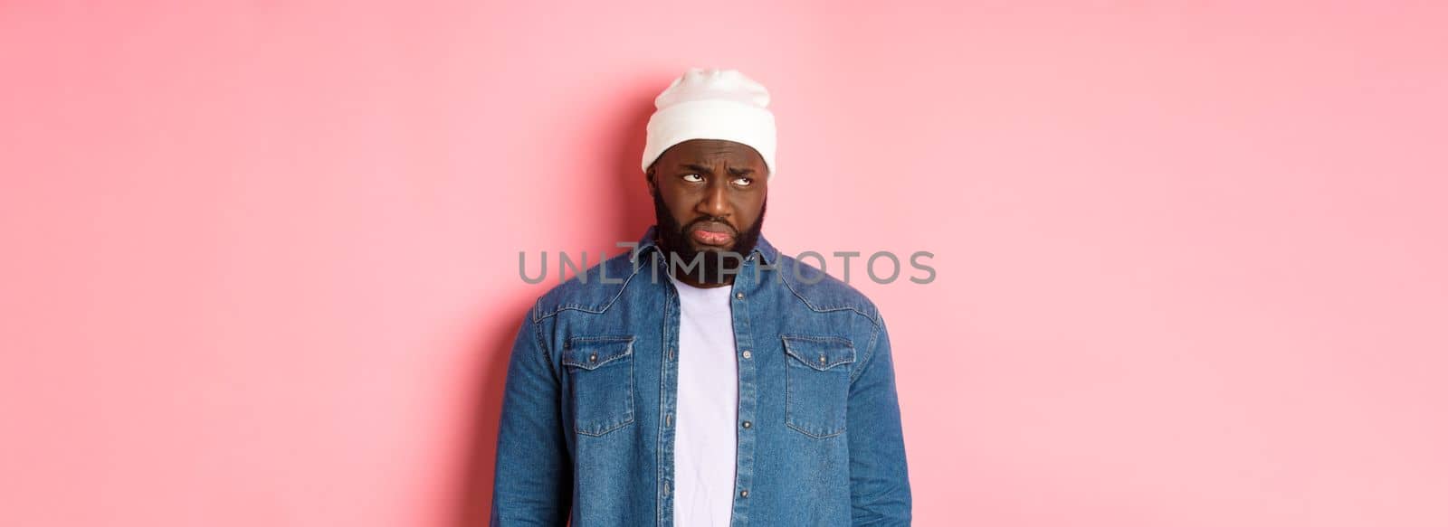 Disappointed and sad Black man, sulking and whining, looking at upper left corner with offended and jealous grimace, standing over pink background.