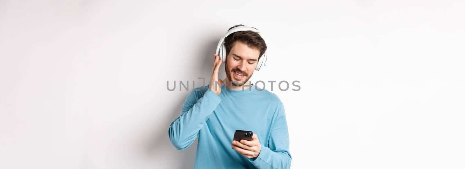 Cheerful smiling man listening music and looking at smartphone, reading message on phone, standing on white background.