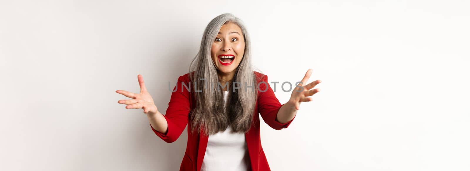 Surprised and friendly asian woman reaching hands forward and smiling, welcoming you, greeting someone, standing over white background.