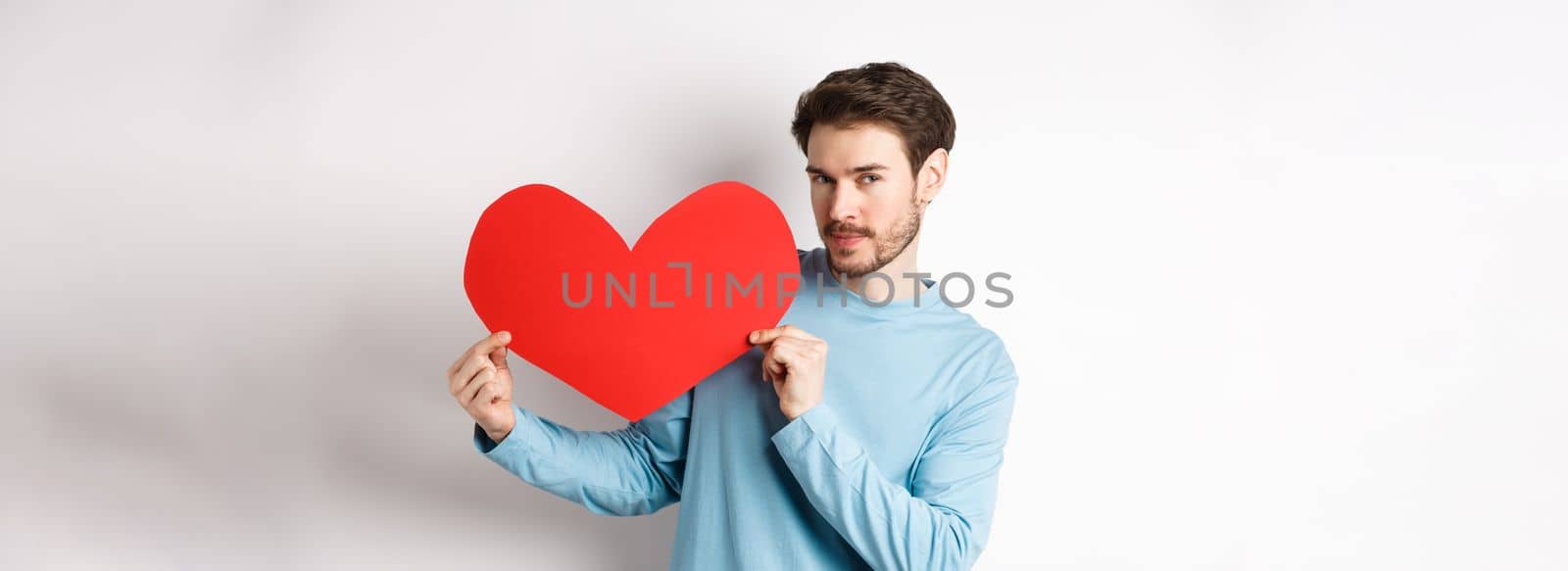 Valentines day. Handsome and romantic man holding big red valentine heart cutout, looking seductive at camera, making love confession, white background by Benzoix