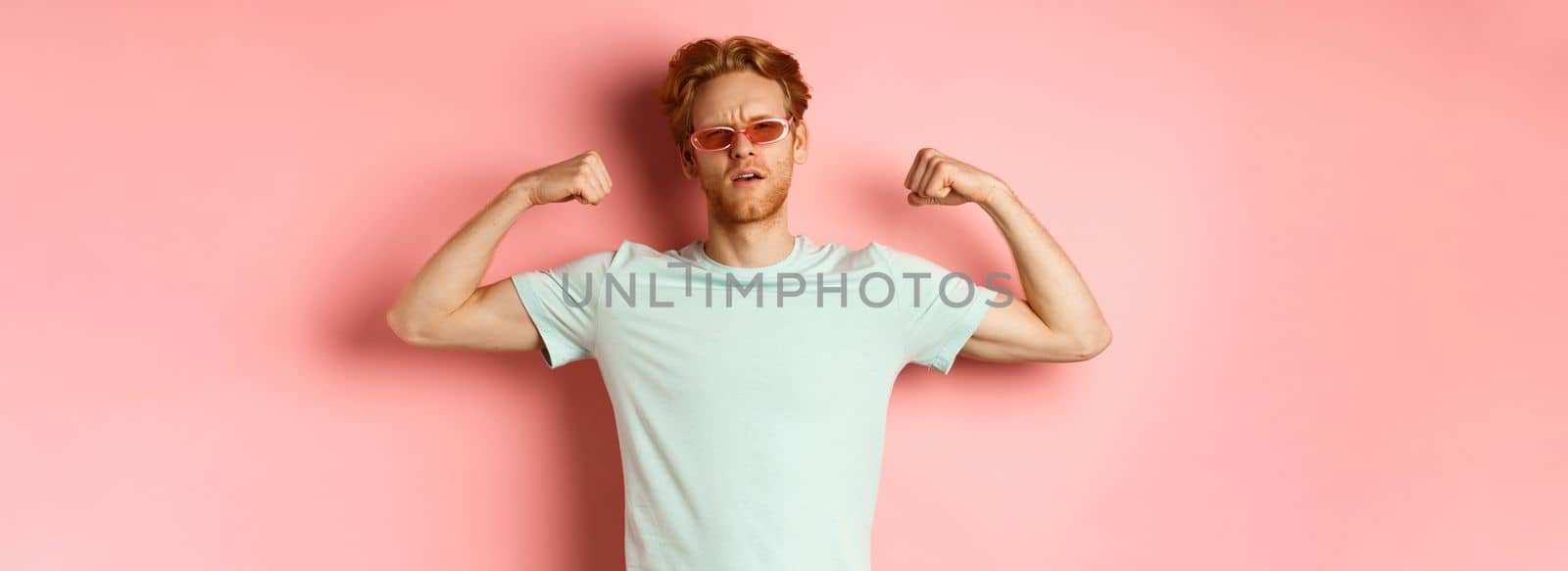 Confident young man with red hair, wearing summer sunglasses and t-shirt, showing strong and fit body muscles, flex biceps and staring cool at camera, pink background. Workout concept