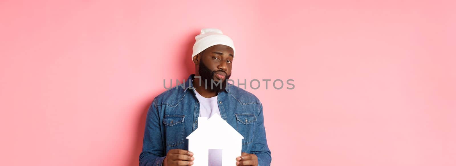 Real estate concept. Skeptical Black male model holding paper house model, looking suspicious and doubtful at camera, searching apartment, standing over pink background.