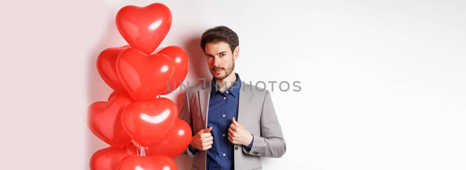 Lovers day. Handsome and confident young man getting dressed for Valentines day, fixing suit and looking at camera, standing near romantic heart balloons, white background.
