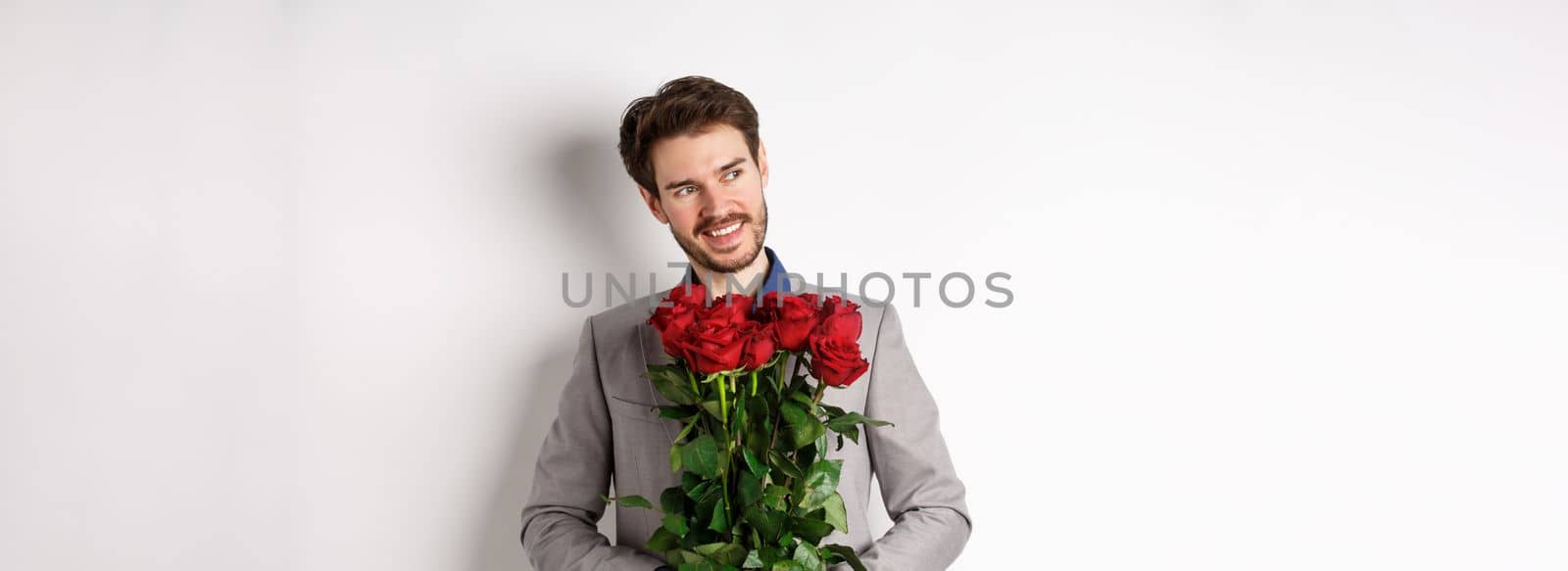 Charming smiling man going on romantic date, wearing suit, holding bouquet of roses and looking left, standing on valentines day with gift on white background by Benzoix