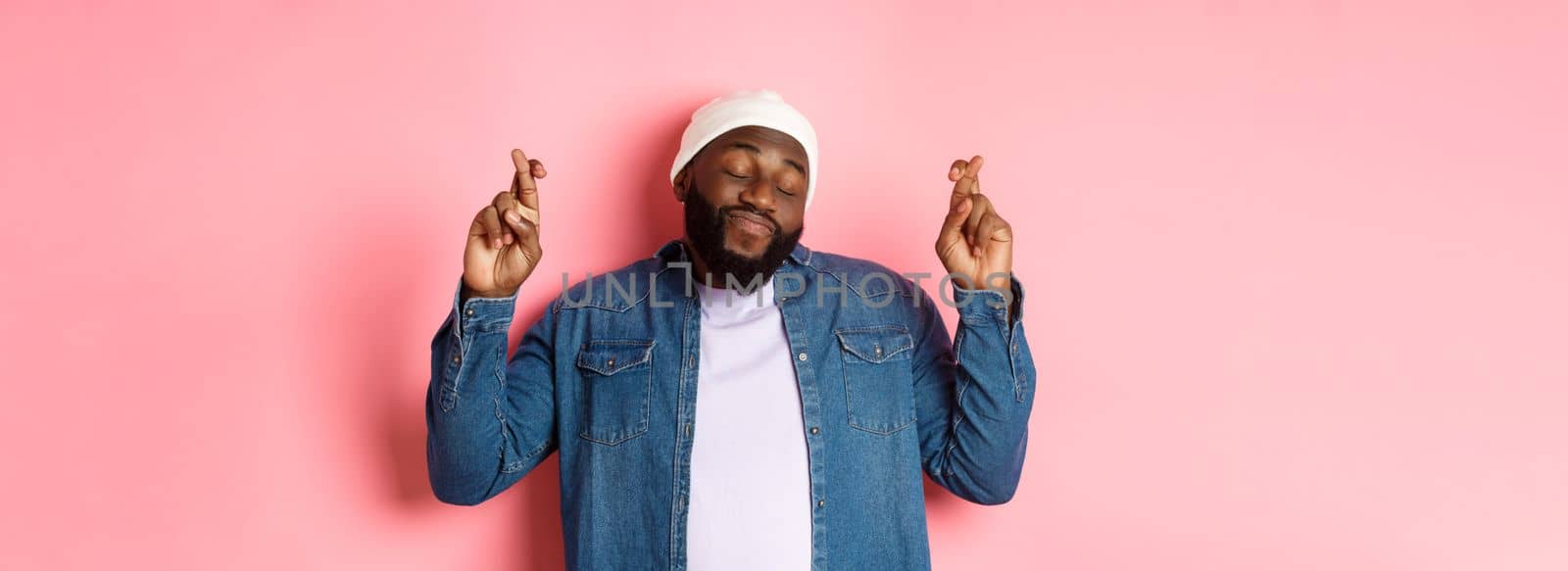 Hopeful and dreamy Black man making wish, cross fingers and smiling, standing over pink background.