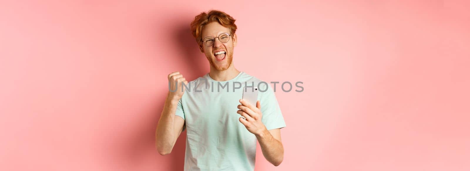 Happy redhead guy in glasses and t-shirt winning online prize, shouting yes with joy and satisfaction, holding smartphone and making fist pump, pink background.