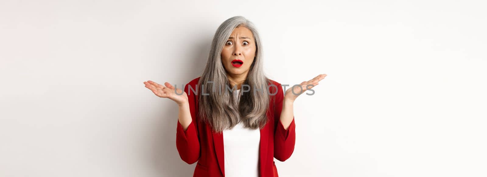 Business people. Confused and anxious asian senior office lady looking worried, spread hands sideways puzzled, standing over white background.