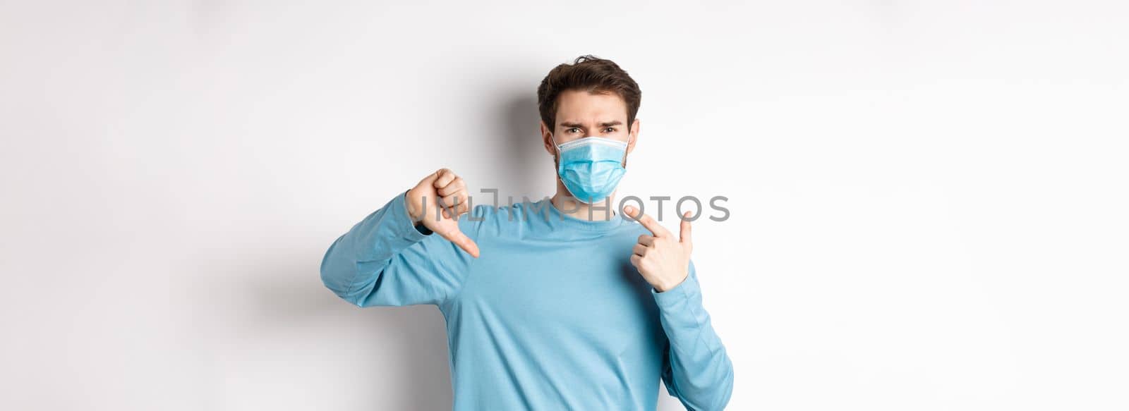Coronavirus, health and quarantine concept. Angry man pointing at his medical mask and showing thumbs down, complaining on covid, white background by Benzoix