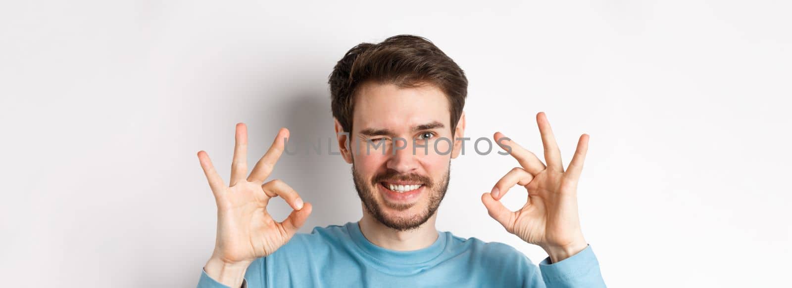 Close up of handsome bearded man winking and smiling, showing okay signs, assure all good, standing on white background by Benzoix