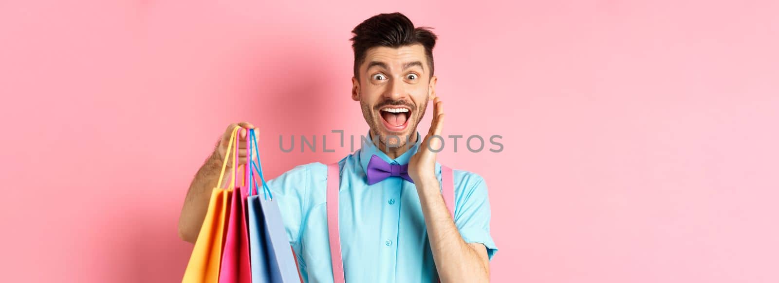 Surprised and happy man screaming of joy and holding shopping bags, shopper seeing discounts, standing over pink background.
