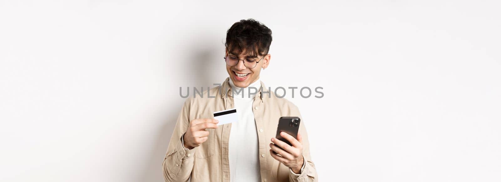 Happy young guy in glasses making online purchase, looking at plastic credit card and holding smartphone, standing on white background.