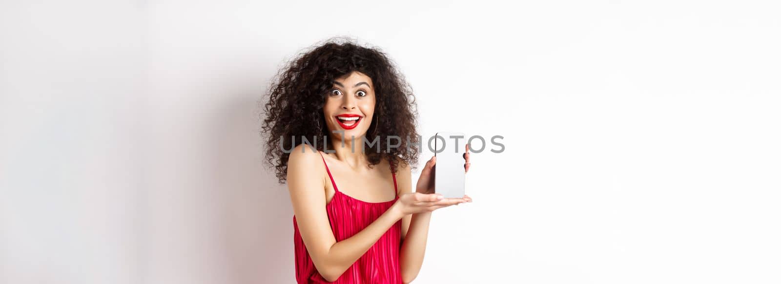 Fashionable lady in red dress and makeup, showing mobile phone screen and smiling, introduce smartphone application, standing over white background.