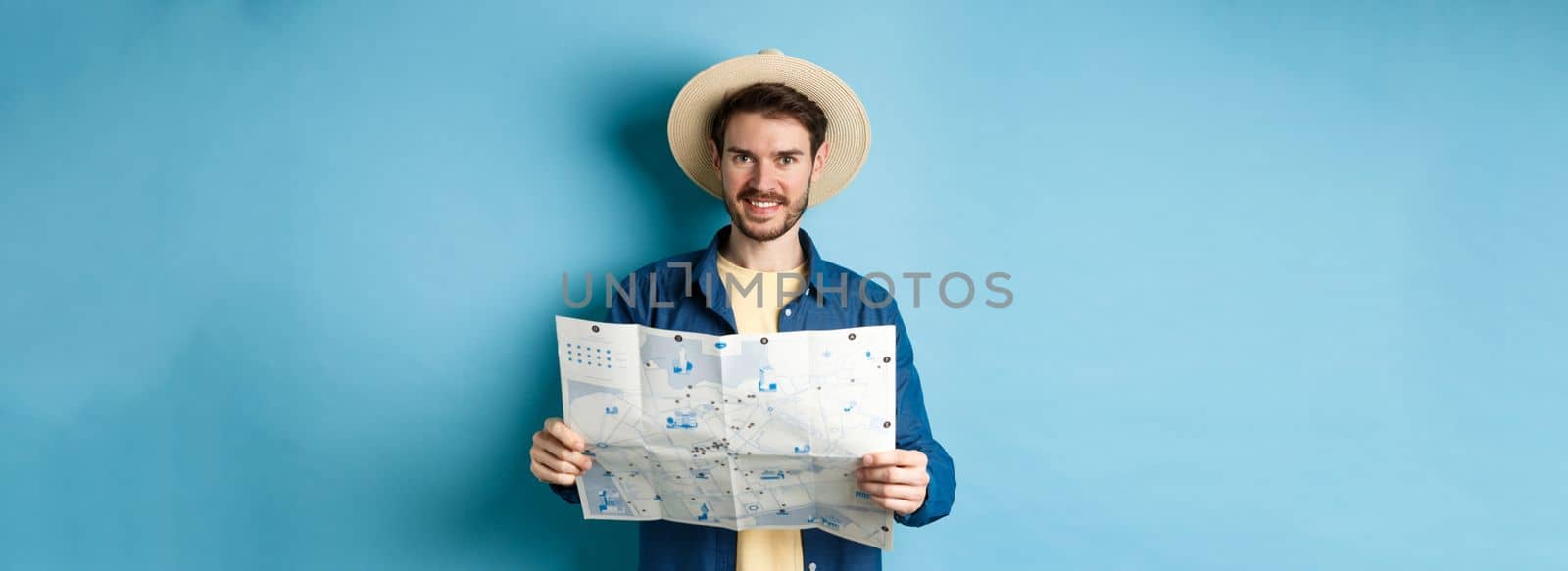 Happy and positive guy on vacation, looking at camera and holding map, smiling excited, going on summer travel, standing on blue background by Benzoix