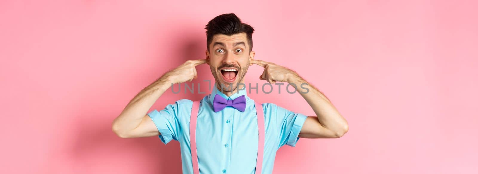 Childish funny guy shut ears and laughing at camera, refuse to listen, standing ignorant on pink background.