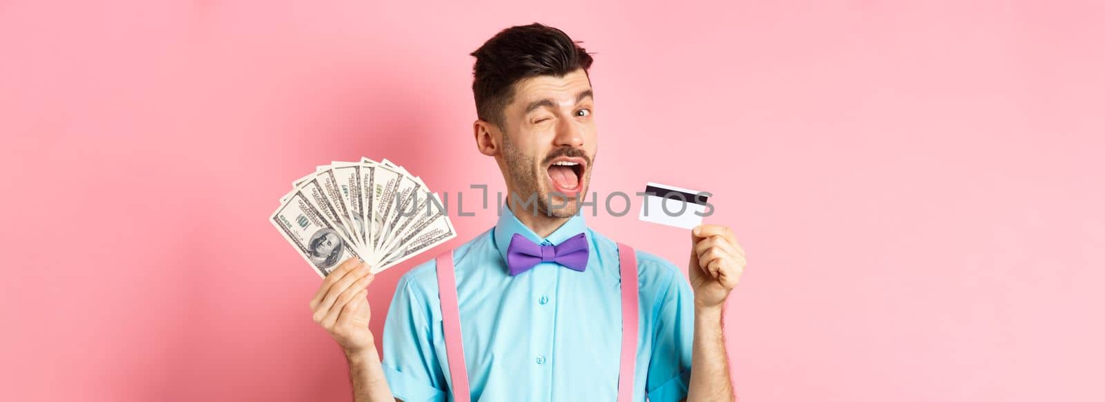Cheerful caucasian man in bow-tie winking at you, showing plastic credit card and money, recommending special deal, standing on pink background.