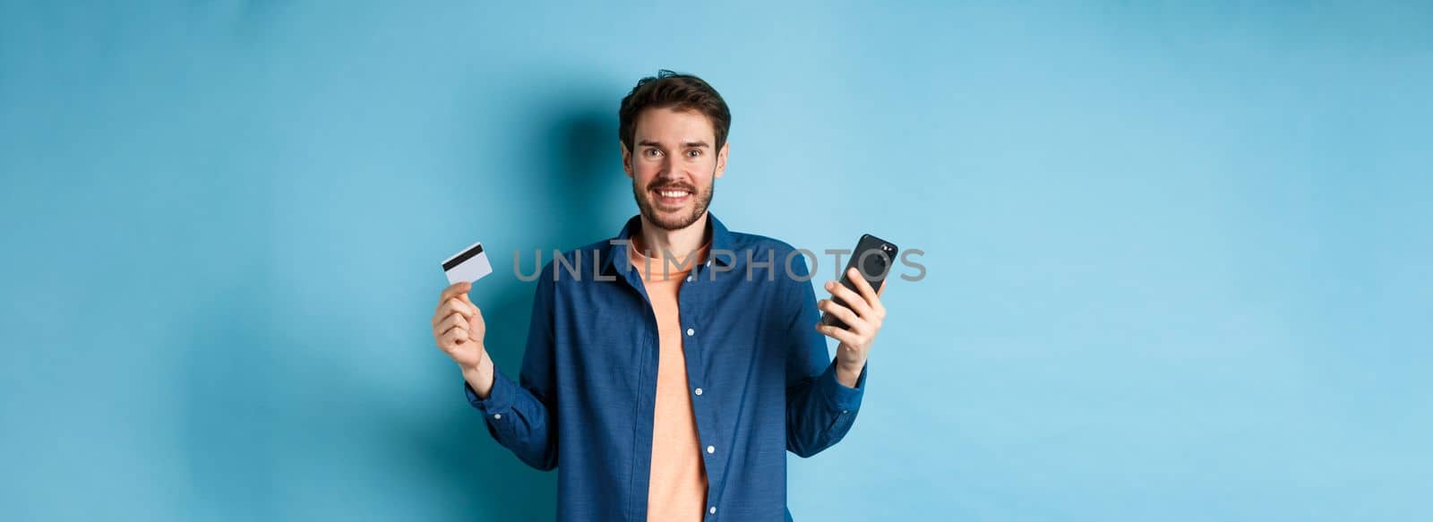 E-commerce concept. Excited young man holding smartphone and plastic credit card, shopping online, standing on blue background.