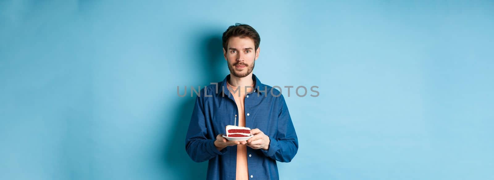 Image of smiling young man celebrating birthday, holding b-day cake with lit candle and looking at camera, standing over blue background by Benzoix