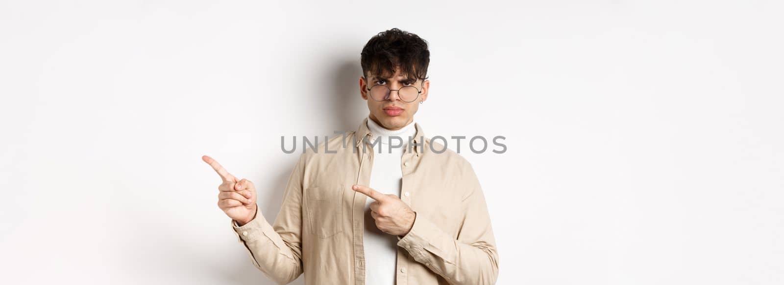 Angry young man frowning and pointing at upper right corner banner, complaining and looking disappointed, standing on white background by Benzoix