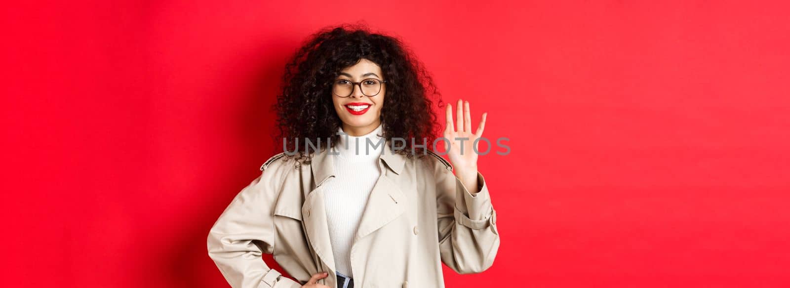 Stylish european woman in glasses and trench coat, waiving hand and smiling, saying hello, greeting someone, standing on red background.