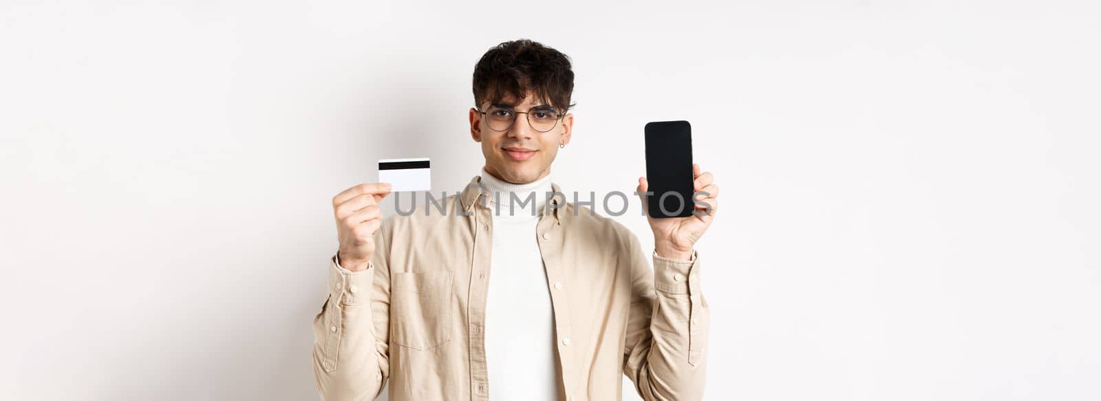 Online shopping. Young modern guy showing plastic credit card and empty smartphone screen, demonstrate account, standing on white background.