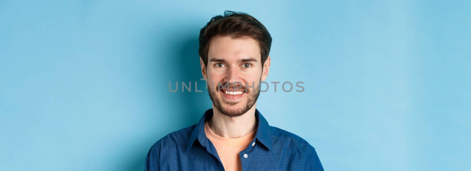 Close up of happy and healthy young man with beard smiling cheerful at camera, standing on blue background. Copy space