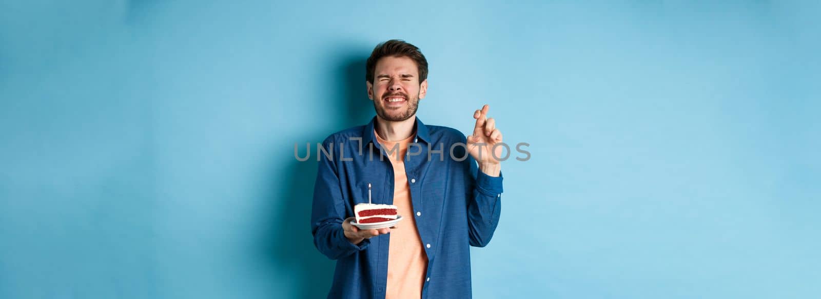 Celebration and holiday concept. Hopeful guy cross fingers and making birthday wish on cake candle, standing on blue background by Benzoix