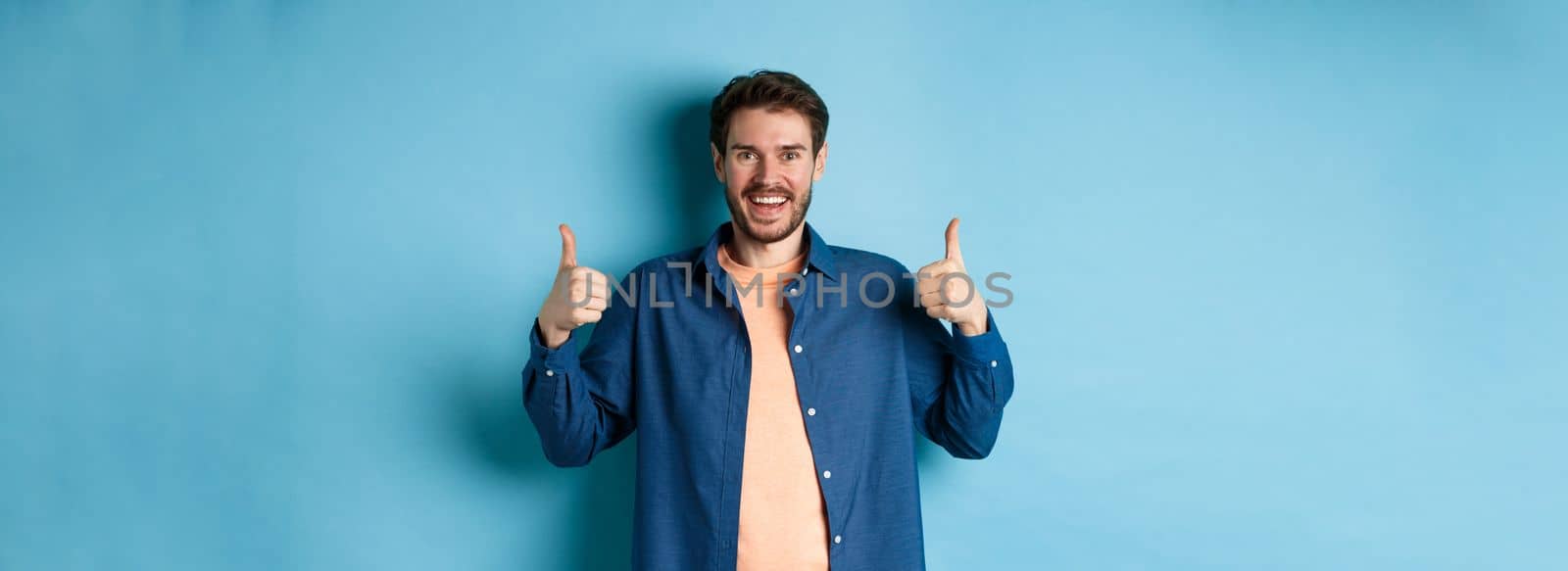Cheerful caucasian guy in casual shirt showing thumbs up in approval, smiling and praising something good, standing on blue background by Benzoix