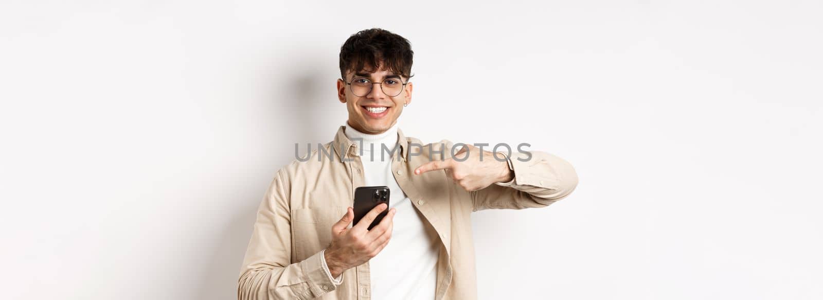 Real people. Handsome young man in glasses pointing finger at smartphone screen, showing online promo, standing on white background by Benzoix