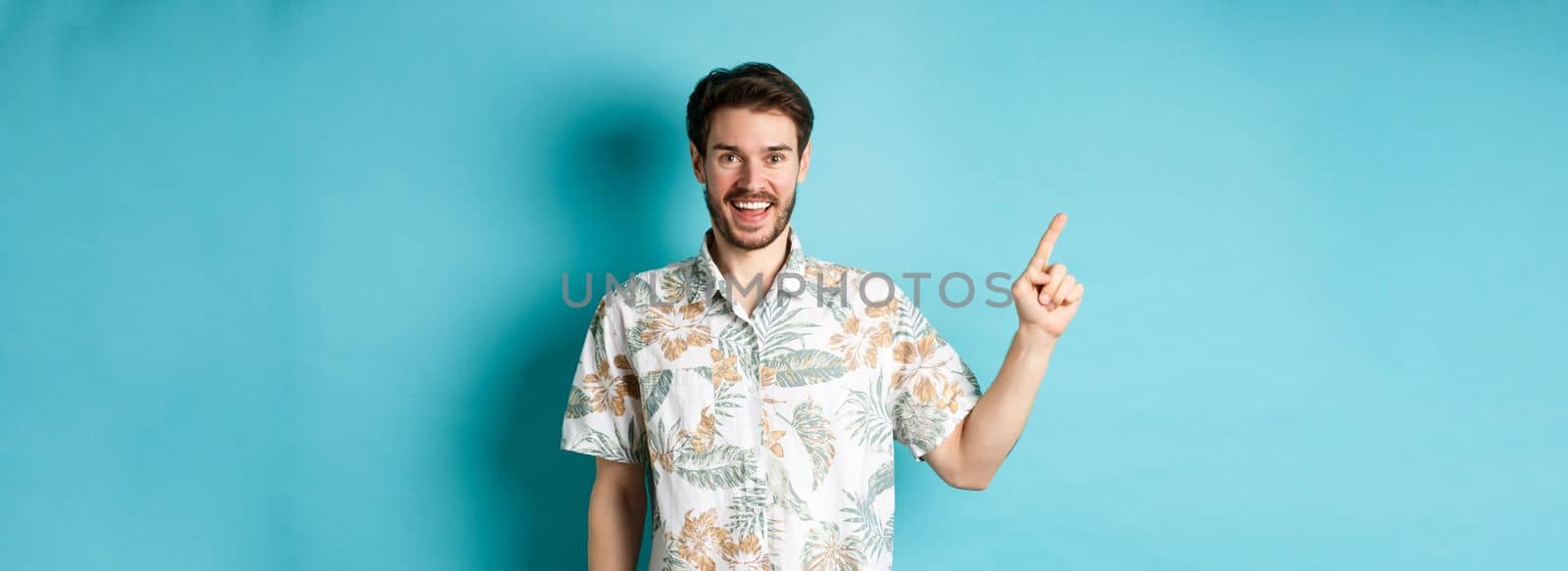 Cheerful young man in hawaiian shirt pointing finger left, showing empty space and smiling. Concept of tourism and vacation.