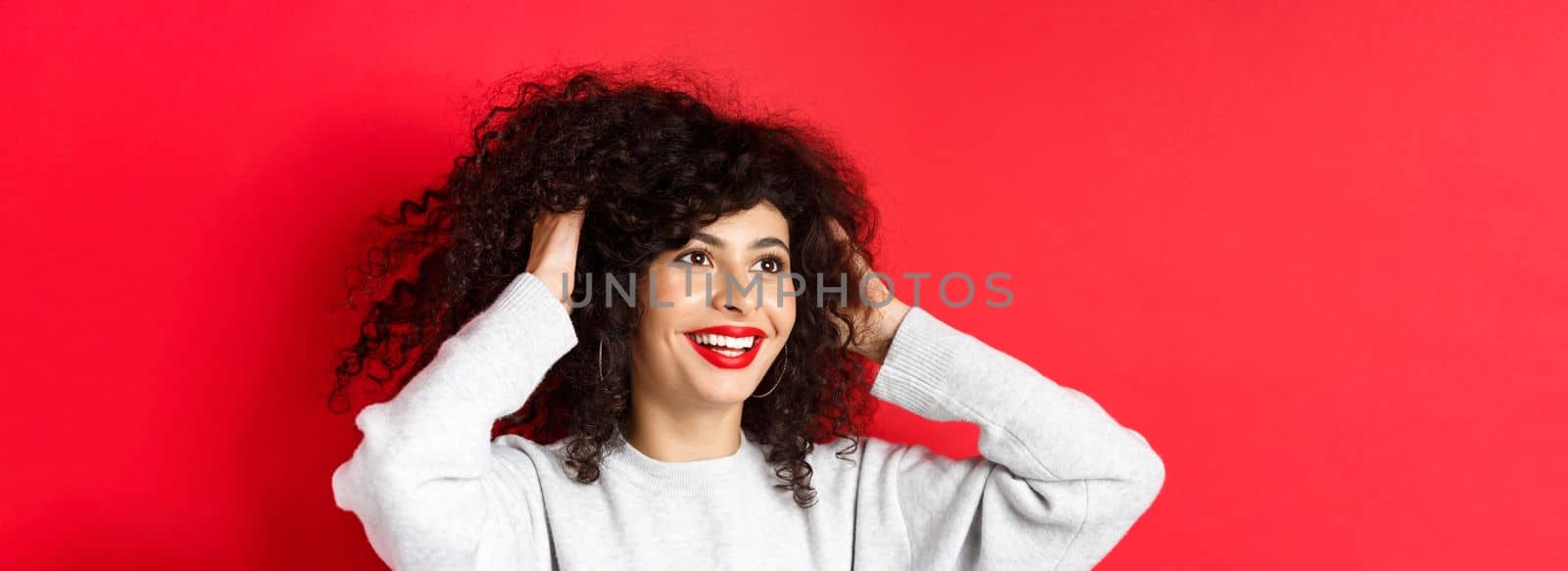 Happy beautiful woman touching natural curly hair and smiling pleased, looking left, like her hairstyle, standing against red background by Benzoix