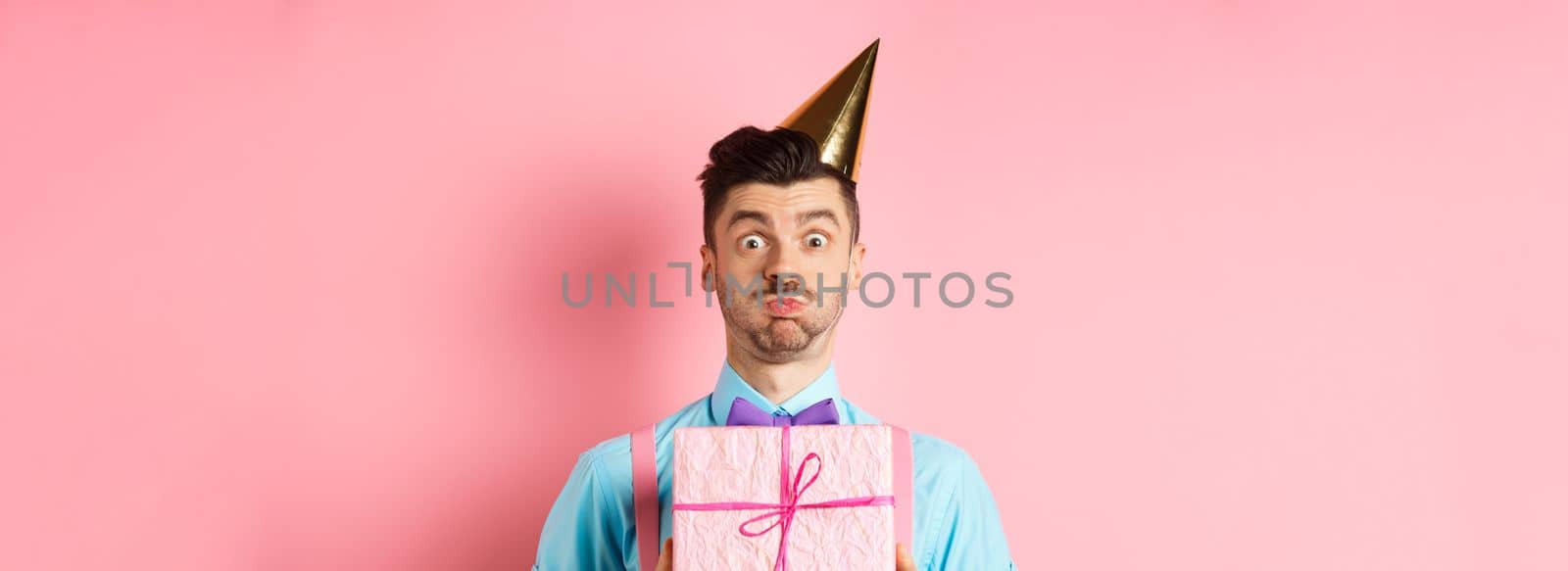 Holidays and celebration concept. Funny guy staring at camera surprised, wearing party hat, holding birthday gift and holding breath, pouting at camera, pink background.
