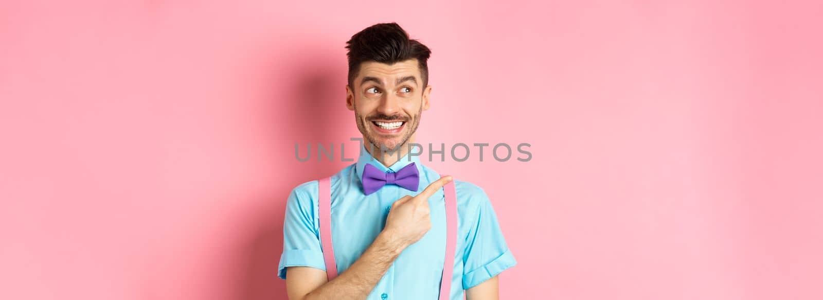 Happy smiling man in funny outfit and bow-tie, pointing finger left and looking at logo, showing advertisement on pink background by Benzoix