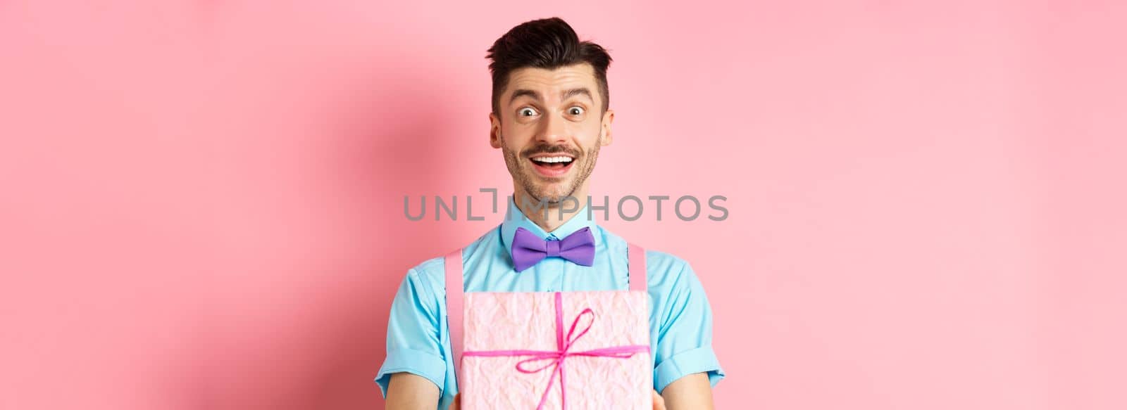 Holidays and celebration concept. Cheerful guy wishing happy birthday and giving you gift wrapped in box, standing over pink background in festive clothes by Benzoix