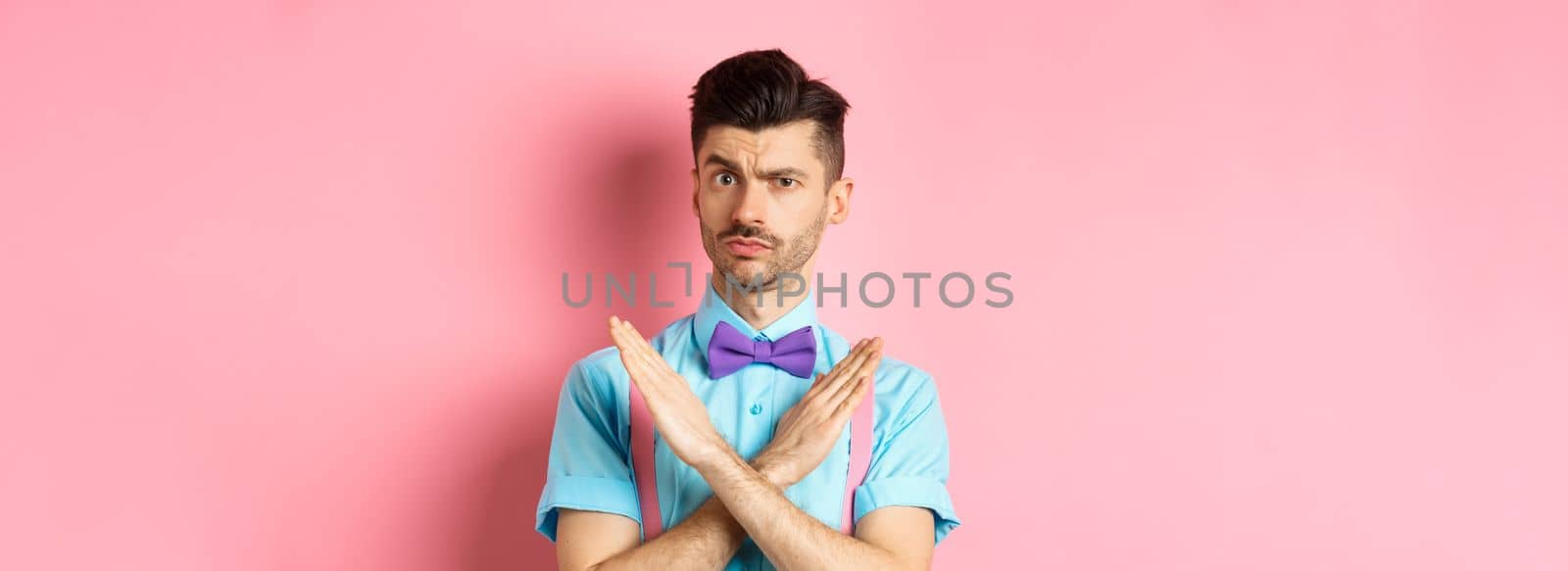 Serious-looking skeptical guy showing cross sign to stop, prohibit something bad, forbid action, standing on pink background by Benzoix