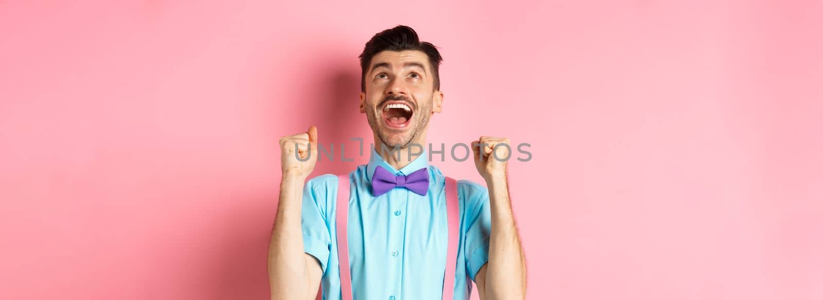Relieved and happy man thanking god, looking up in sky while celebrating victory, winning prize and cheering, triumphing over pink background by Benzoix