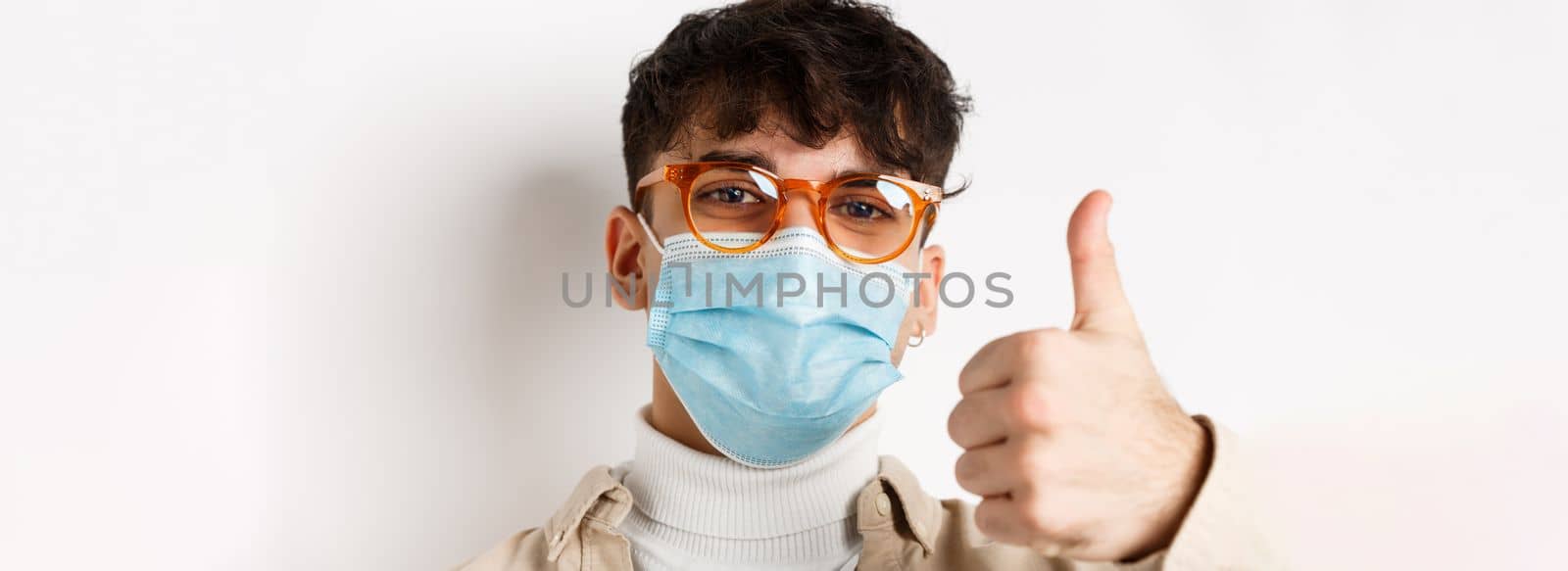 Portrait of young man in glasses and medical mask showing thumb up, smiling with eyes, standing on white background. Covid-19 and pandemic concept.