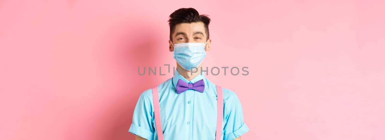 Covid-19, pandemic and health concept. Portrait of smiling man in medical mask feeling happy, standing in festive bow-tie and suspenders, pink background by Benzoix