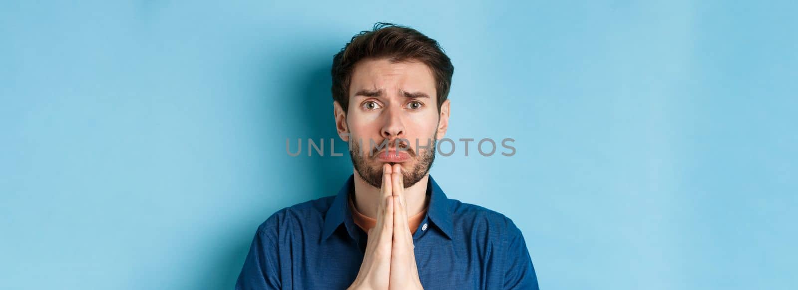 Sad young man begging for help, apoligizing and sobbing miserable, standing on blue background.