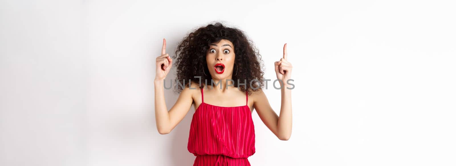 Impressed young woman with curly hair, wearing red dress, gasping and saying wow, pointing fingers up at logo, standing over white background.