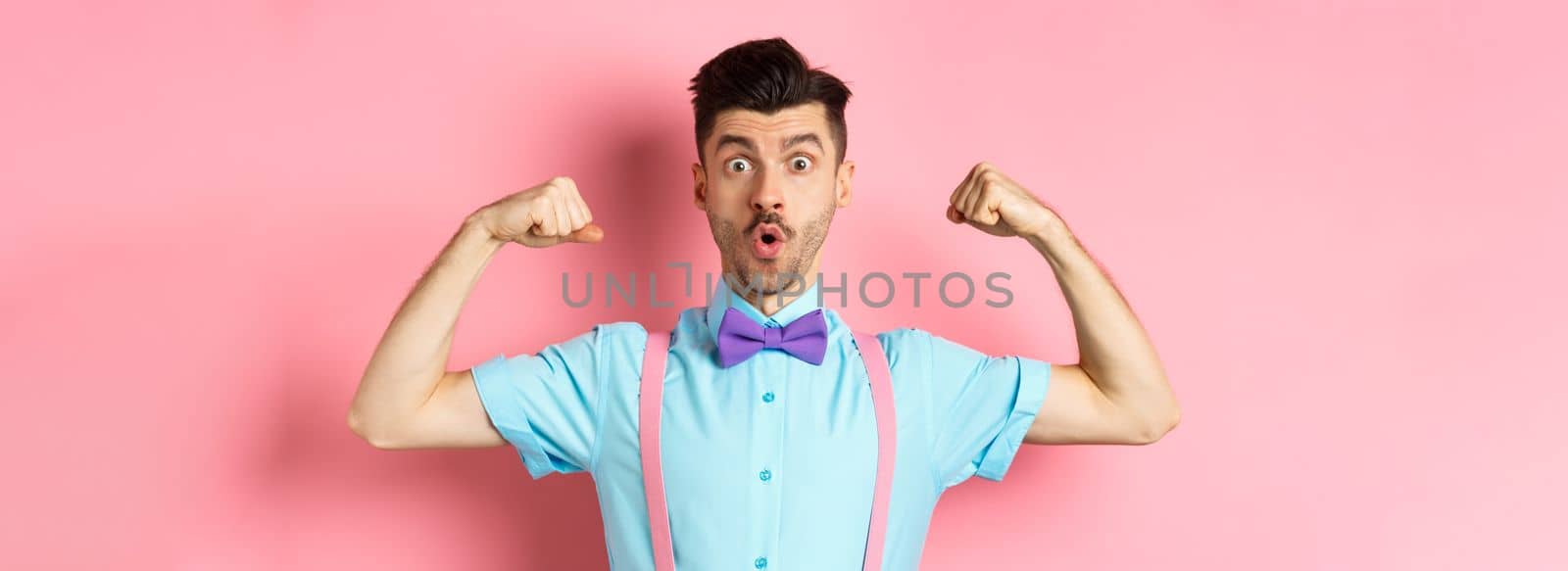 Image of funny guy with moustache and bow-tie, showing muscles, flexing biceps and looking surprised at camera, standing in disbelief on pink background by Benzoix