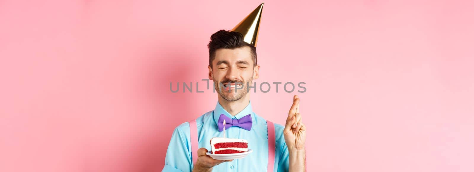 Holidays and celebration concept. Hopeful birthday guy in party hat making wish, holding cake with candle and cross fingers for dream come true, pink background by Benzoix