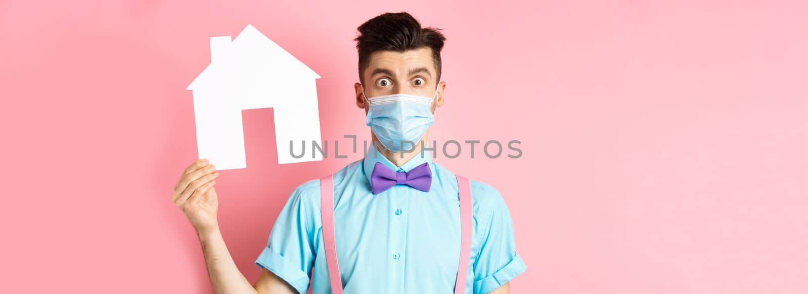 Covid, pandemic and real estate concept. Confused guy in medical mask searching property, showing house cutout and stare at camera worried, standing over pink background by Benzoix