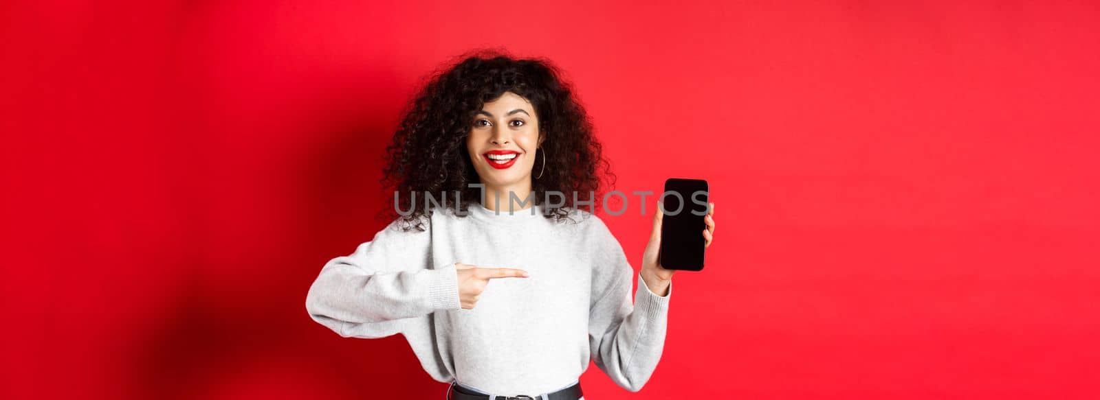 Beautiful female model showing empty smartphone screen, pointing at phone display and smiling, standing against red background.
