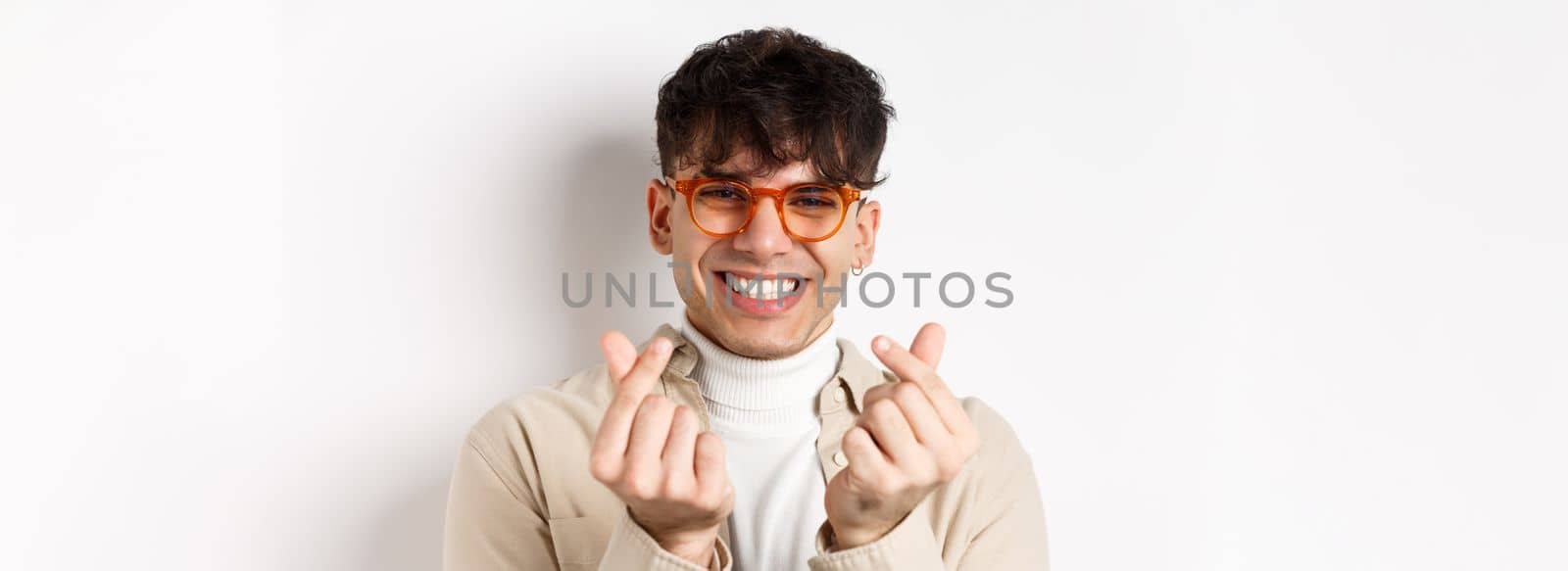 Cute young guy in glasses smiling and showing finger hearts, standing on white background by Benzoix