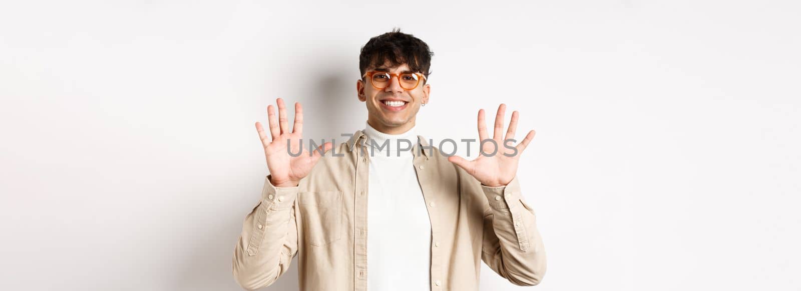 Modern guy in glasses and stylish outfit, showing ten fingers number and smiling, standing on white background by Benzoix