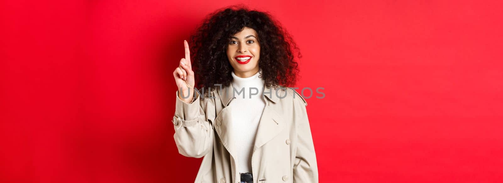 Attractive modern woman with red lips, curly hairstyle, wearing spring trench coat, pointing finger up and smiling, standing against red background by Benzoix