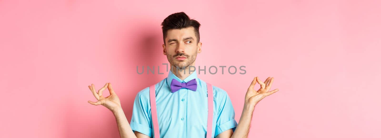 Funny guy with moustache and bow-tie fake meditating, peeking aside while doing yoga asana, standing over pink background by Benzoix