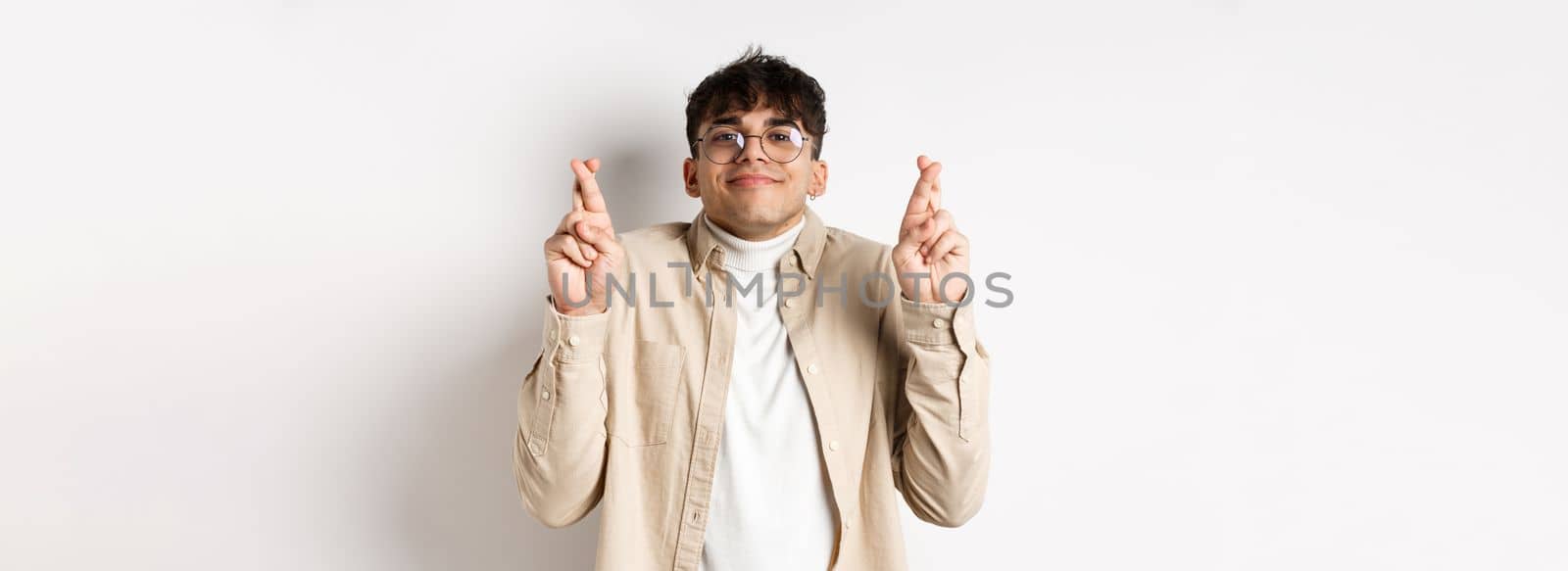 Hope. Happy young man making wish and smiling, looking hopeful at camera as waiting for dream come true, cross fingers for good luck, standing on white background.