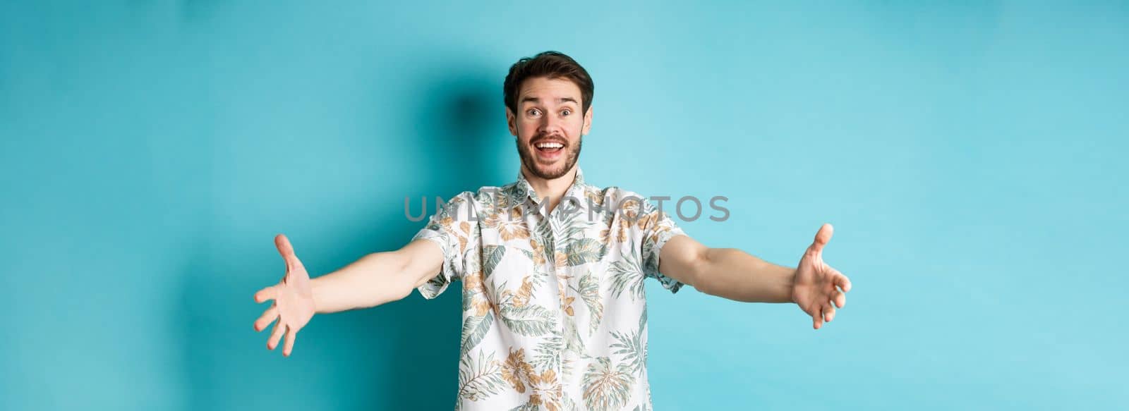 Summer holiday. Cheerful tourist stretch out hands for hug, welcome someone with friendly smile, greeting you, standing on blue background.